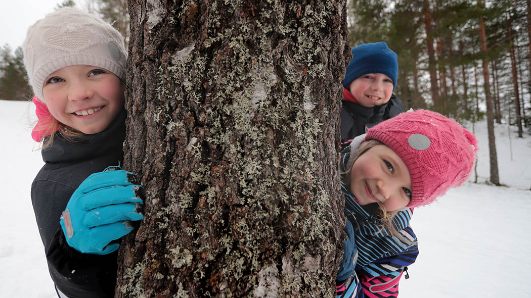 Children peeking out from behind a tree in a snowy forest.