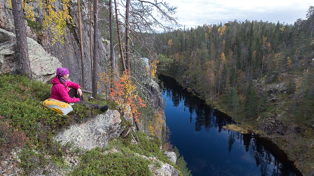 The hiker sits on top of a rock wall and looks at the opening gorge landscape, with an elongated lake at the bottom.