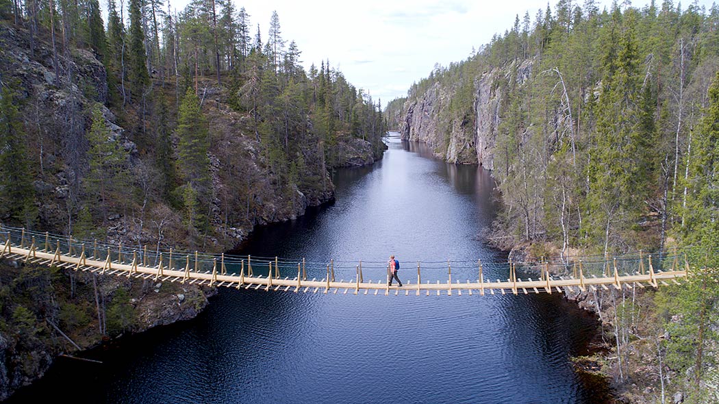 A hiker walks across a suspension bridge over a ravine lake. The bridge is about 10-15 meters above the water. Rocky heights rise at the shores.