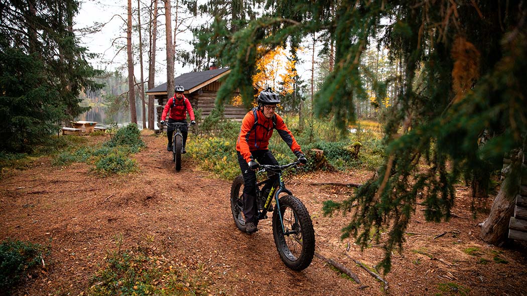Two mountain bikers leaving the yard in autumn weather. A lake and a log building in the background.