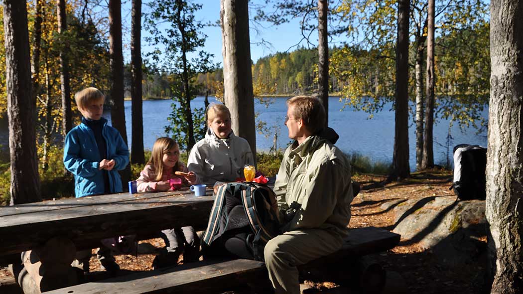 A family at a group of tables in an autumn forest landscape by a lake.