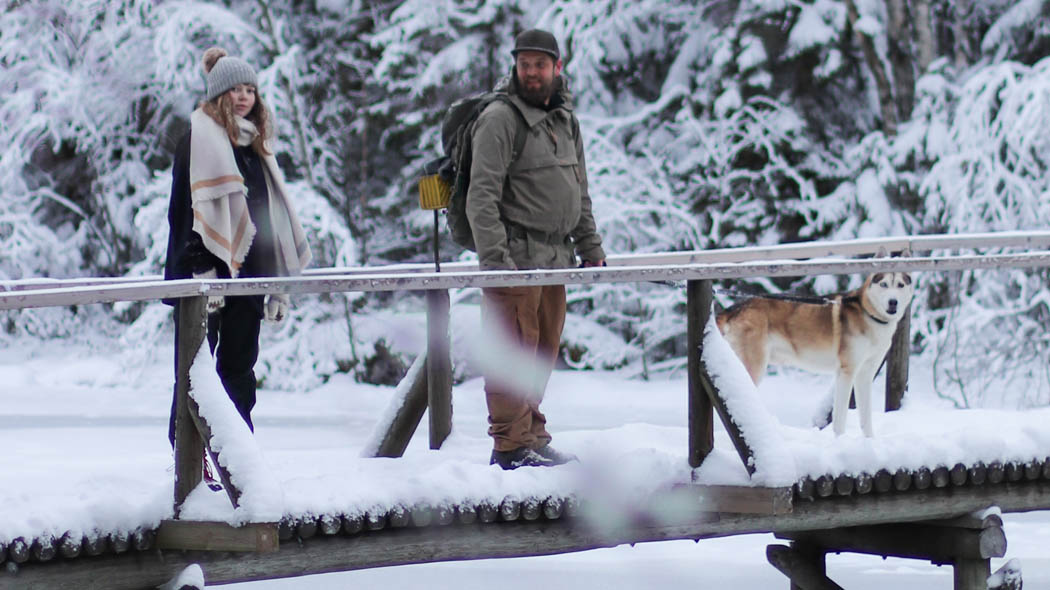 Kaksi retkeilijää ja koira lumisella sillalla.