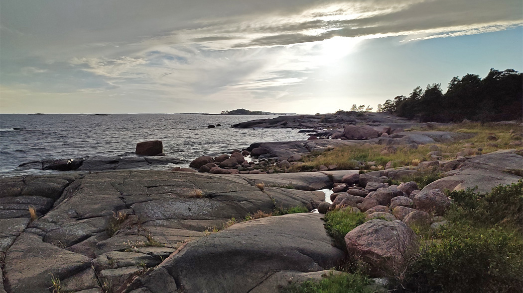 Archipelago landscape.  There is rock in the foreground. In the background there are a coastal forest, a rocky seashore and the horizon. The sun is behind a cloud.