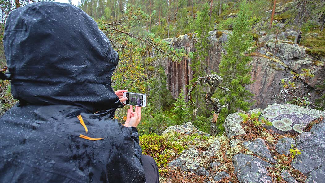 A hiker takes a picture of a rock-walled gorge with a mobile phone in rainy weather. In the foreground, lichen-like rocks and a gnarled tree.