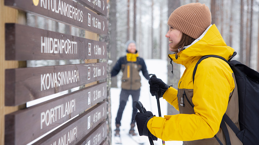 A smiling person reads a signpost. In the background a man and a snowy forest.