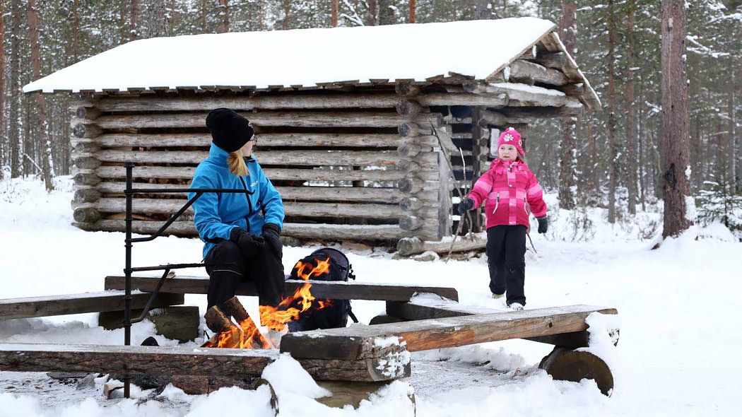 An adult and a child spend time by a campfire in a snowy forest.