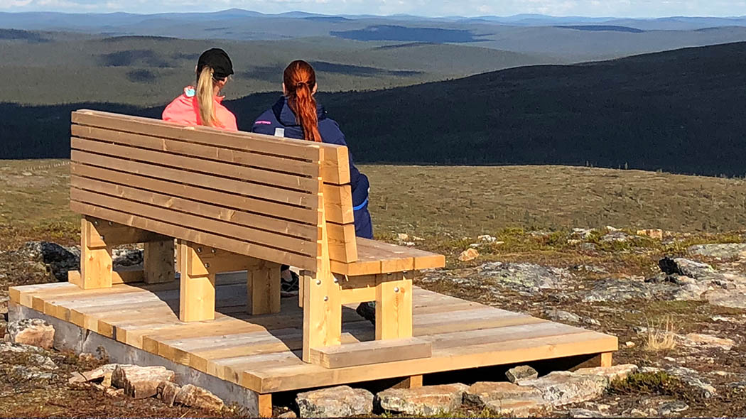 A sturdy bench on top of a fell. Two women are sitting on the bench, admiring the view of the fell landscape.