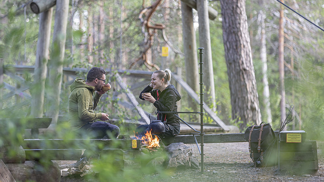 Two hikers are sitting at the campfire site with wooden “Kuksa” cup in their hands.