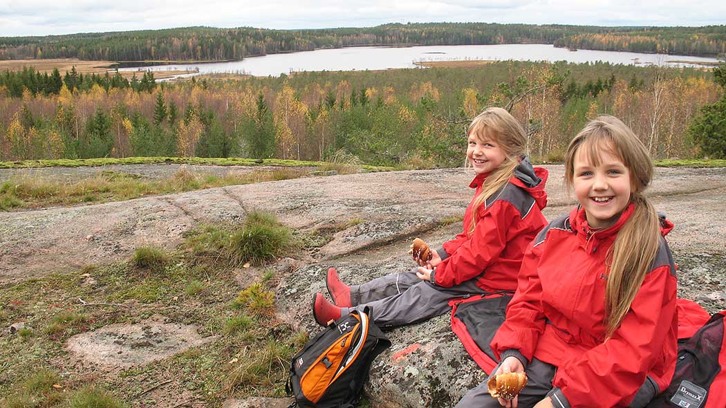 Two smiling children sitting on a rock with bread in their hands. An autumn sea- and forest landscape opens up in the background.