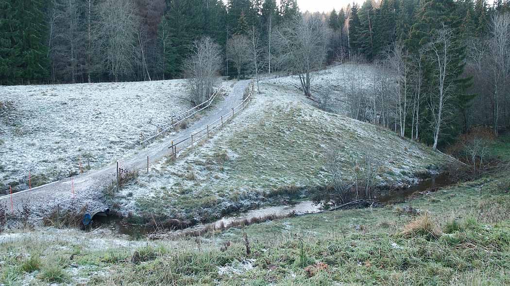 The stream flows across the pasture landscape with little snow. In the middle of the landscape there is a path towards the forest in the background.