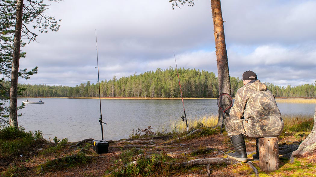 A fisherman is sitting on a wooden plank on the shore of a lake. Two fishing rods are standing on the shore and the lures are in the water. In the background there is a boat with two people sitting on it.