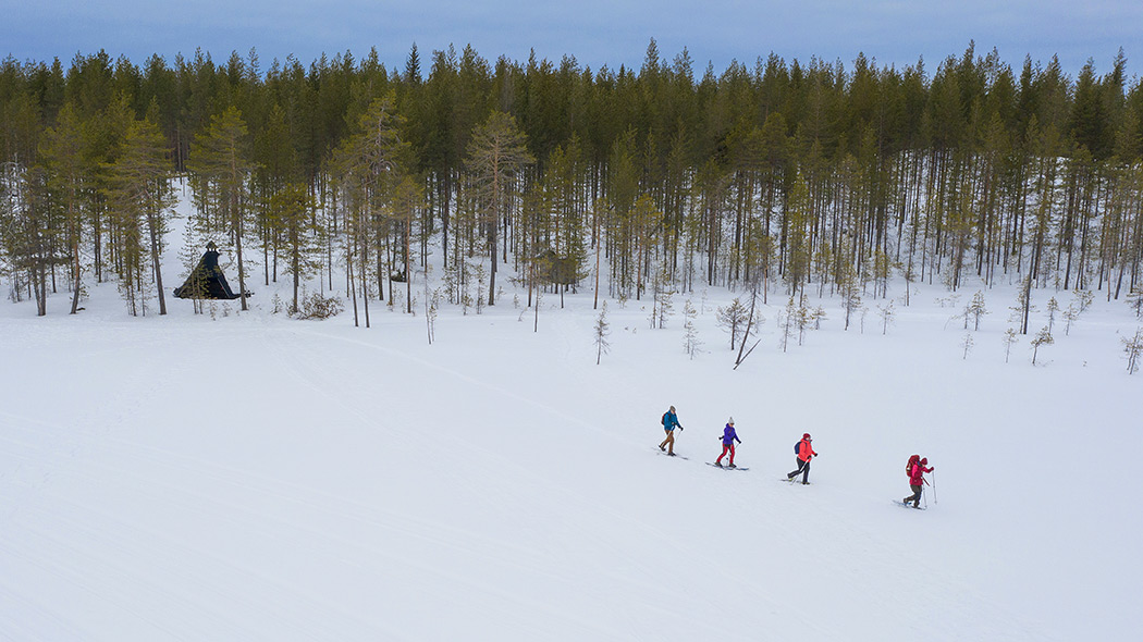 Four snowshoers in the aerial view are walking from a log cabin across a snowy pond.