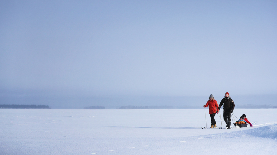Two adults ski on a snowy and open lake. The other is followed by a crib with two children.
