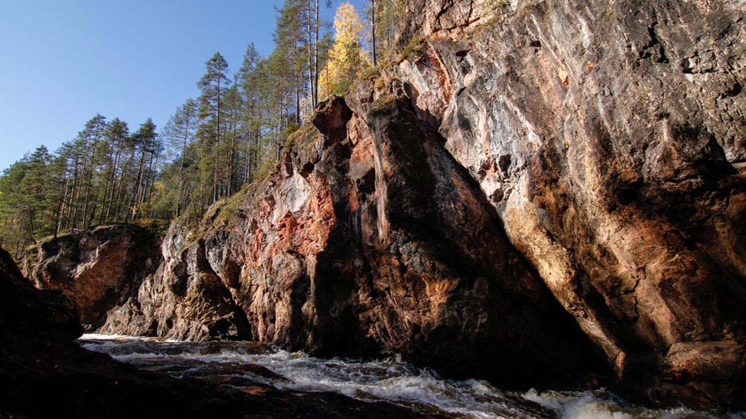 A landscape picture where a dolomite rock rises upwards and below the picture you can see a rapid current. There are trees on top of the rock.