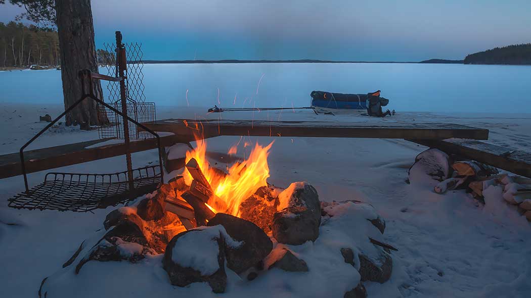 Campfire by the lake. Behind the campfire are benches and a snow-covered lake. There is snow on the ground.