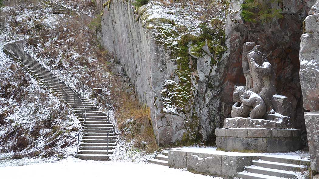 A steep cliff with a bear family statue at the base. There are stone stairs on the slope. There is a little snow on the ground.