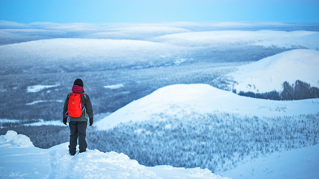 A woman with a red backpack is standing on a snowy fell. Several fells can be seen on the horizon. There is snow everywhere.