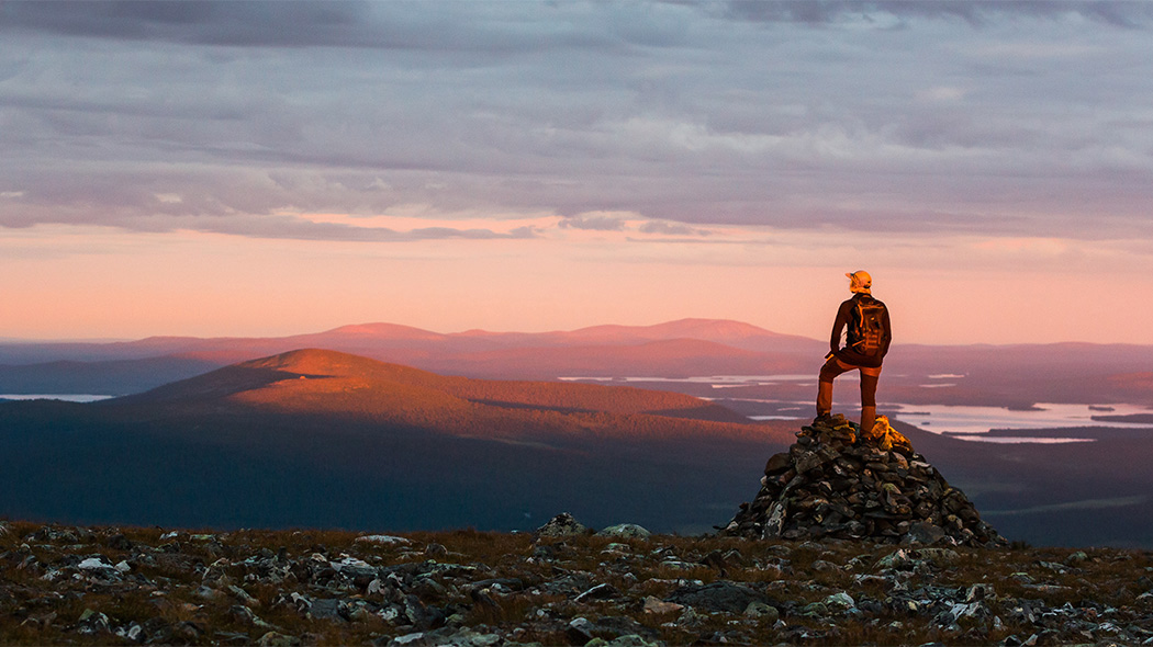 A hiker stands on an open fell top and looks down at the landscape. Several fells can be seen on the horizon. It's a summer night.