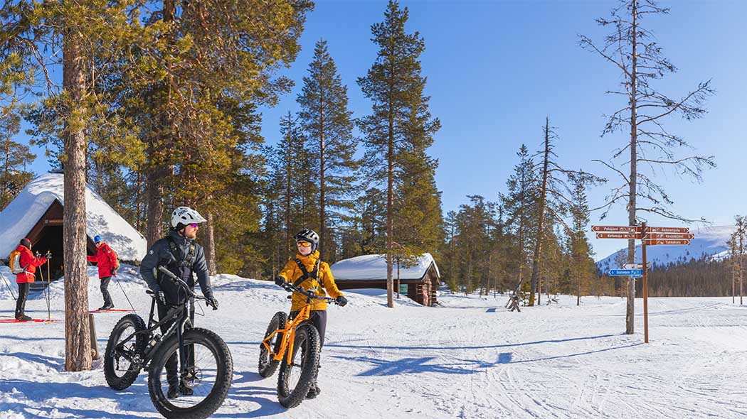 Two people standing in the snow, leaning against black fat bikes in the foreground. There is a lapp-hut and a woodshed in the background and pine forest and a blue sky can be seen in the wintry landscape.