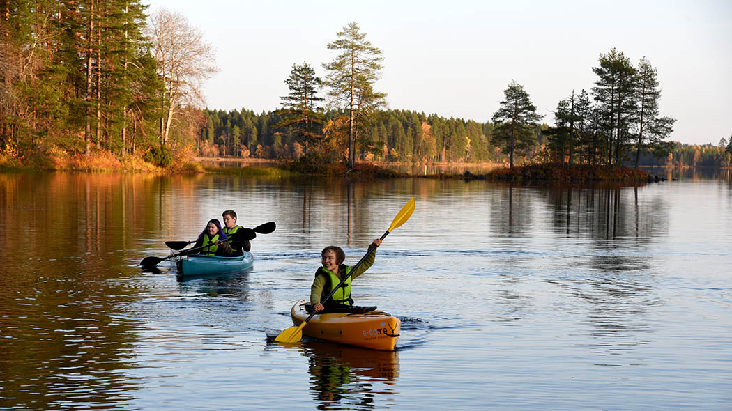 A lone kayaker in the foreground and a double kayak with two paddlers in the background. Forest in autumn colours can be seen on the shore.