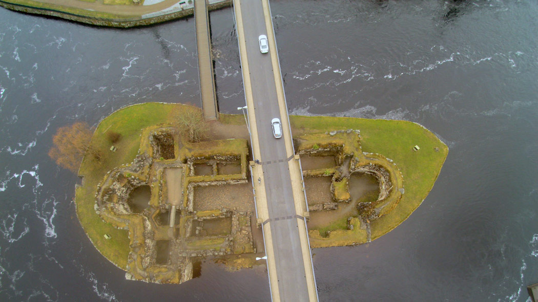 A castle ruin on a small island photographed from above. There is a road leading across the river and the castle.