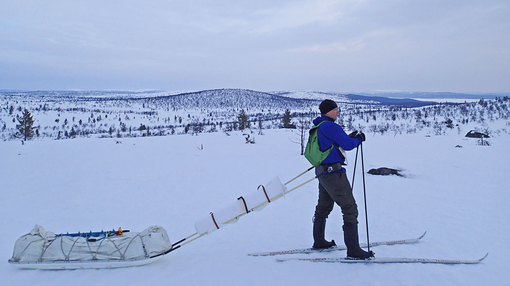 Snow covers the fells and here and there glimpses of blue slopes of hills. A man pictured sideways with a sledgehammer with an ice auger on top, a man with skis on his feet.