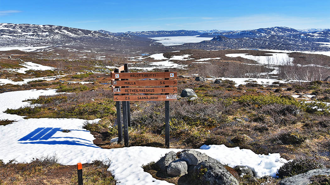 The picture shows open fell terrain, where there is still a lot of snow. In the middle of the picture are wooden signposts that say e.g. The Nordkalott Trail
