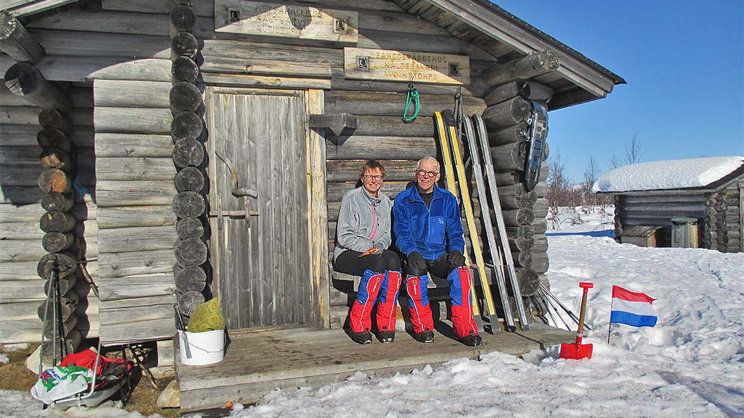 A man and a woman are sitting on the porch of a small log cabin in the winter. They have ski equipment and skis upright on the wall and a small French flag in the snow. In the background is a small wooden outbuilding.