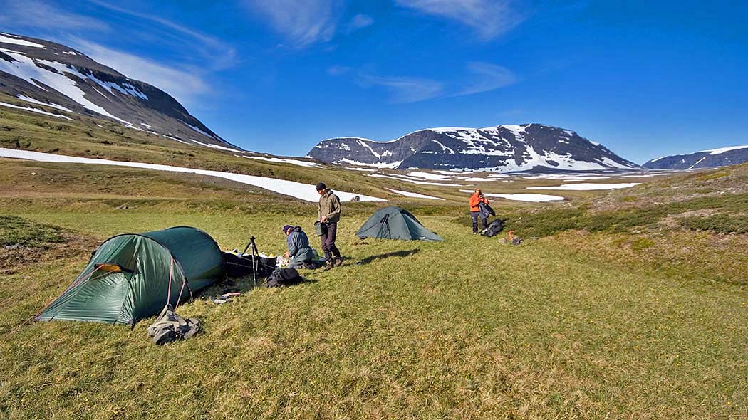 Three people setting up tents in open fell area in sunny weather. High fell tops in the background.