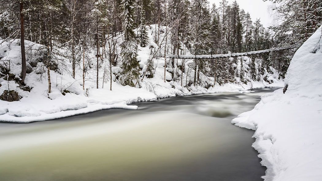 A snowy landscape with a suspension bridge above a flowing river.