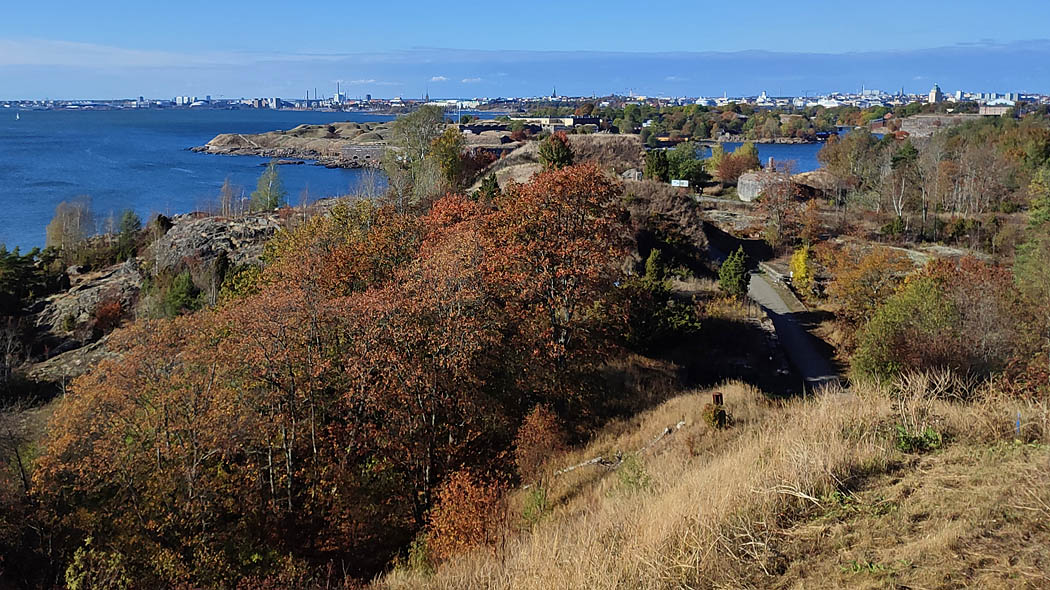 Autumn landscape from a high grassy slope, in the foreground trees, shrubs and open rock, as well as a narrow road. In the background sea, the city's tall houses, towers and chimneys.