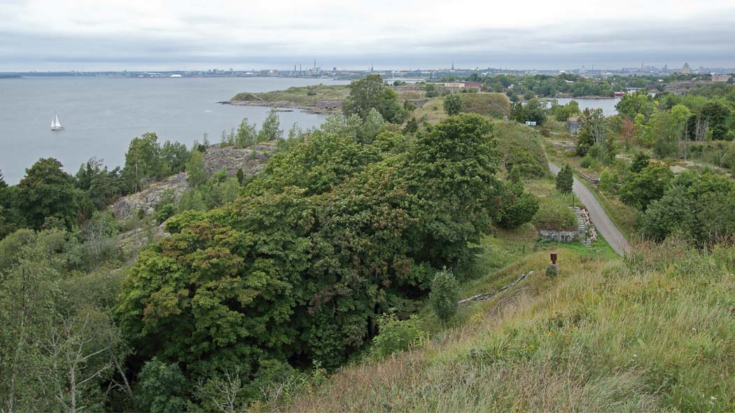 Landscape from a high grassy slope, in the foreground trees, shrubs and open rock, as well as a narrow road and stone stairs. In the background sea, the city's tall houses, towers and chimneys.