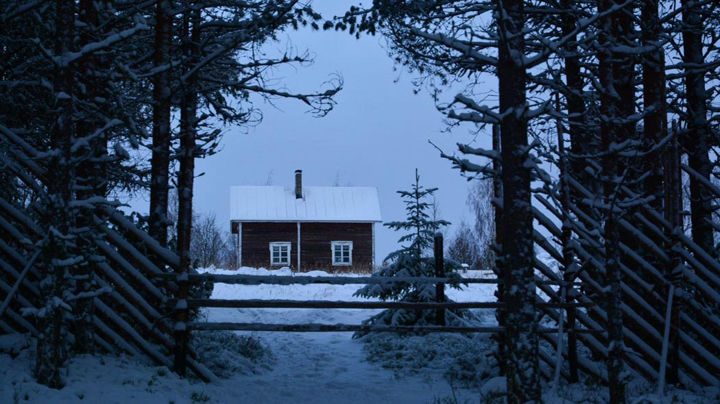 Riukuaidan veräjän takaa näkyy vaaran laella oleva talo. Maassa, puissa ja talon katolla on lunta.
