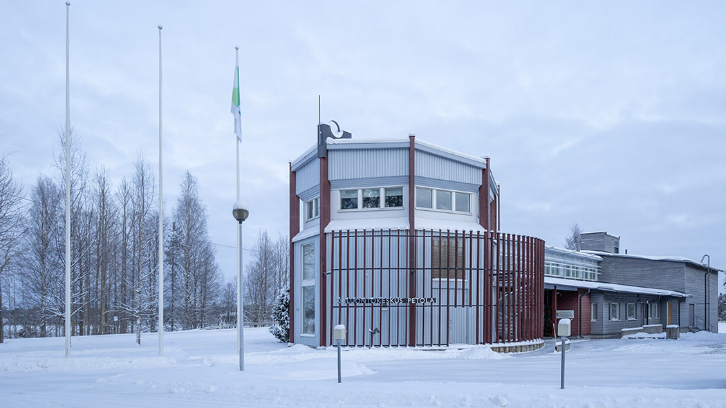 A large building in an winter landscape. In the foreground, three flagpoles, one of which have a flag. There is a snowy lake in the background.