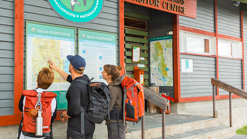The emblem of the national park, an information board and a map can be seen on the outer wall next to the front door of the nature centre. Three hikers are studying the map.