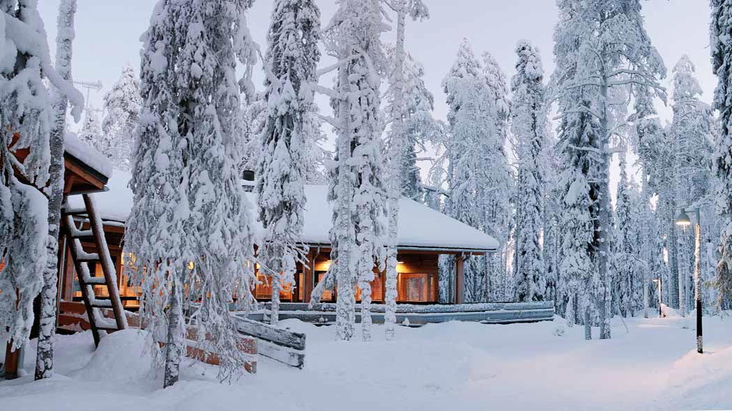The log house, whose interior is lit, stands amidst large trees. The snow is thick on the ground and in the trees.