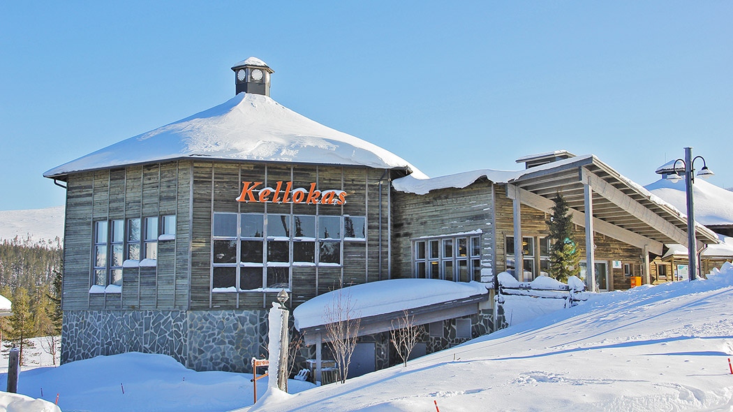In the picture, there is a large modern building with the name Kellokas written on the wall. The building is on a slope. A snowy fell in the background. It's a sunny winter day.