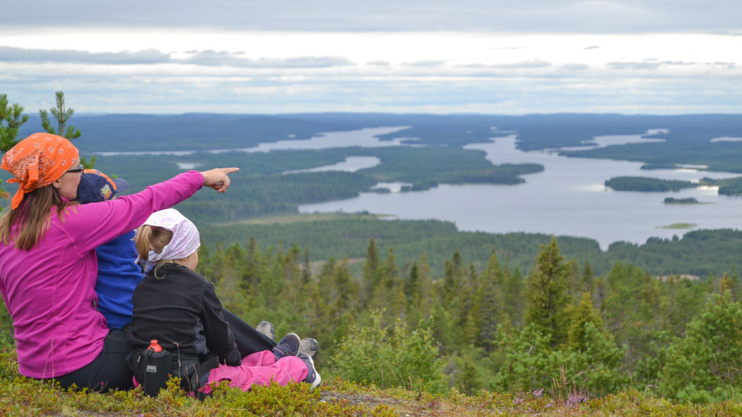 A hiker with children in the lap sitting on a fell slope pointing at the horizon. A forest and lake landscape opens up below.