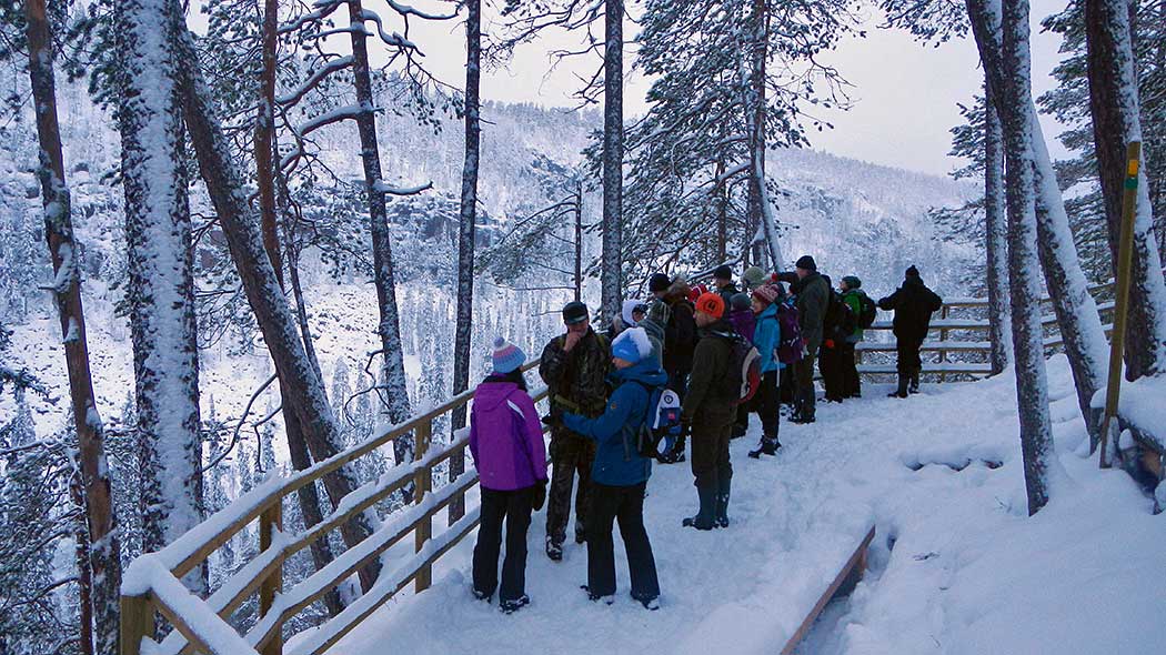People stand at a viewpoint with a railing in winter. In the background is a wide canyon valley.