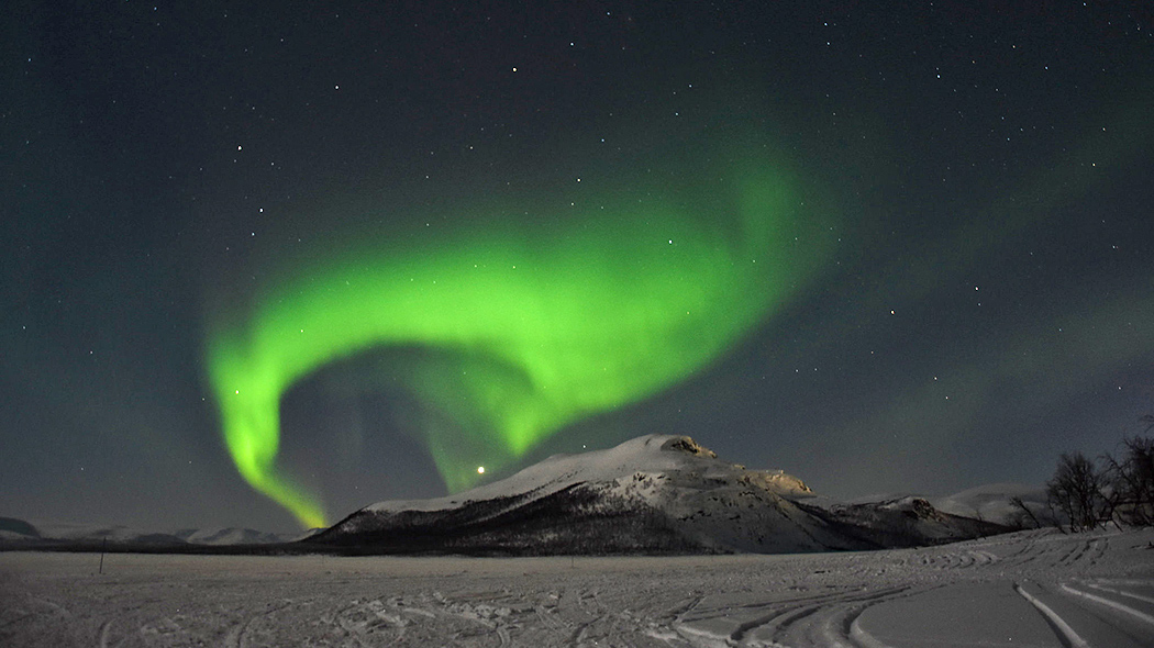 In the foreground a frozen lake, in the background a mountain slope covered with snow. Northern Lights on the dark sky.