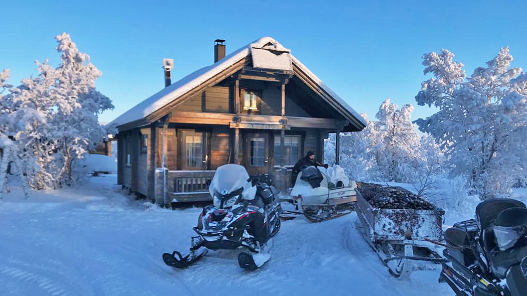 A double storey log cabin in winter seen from the front. There are two snowmobiles in front of the cabin, followed by sleighs.