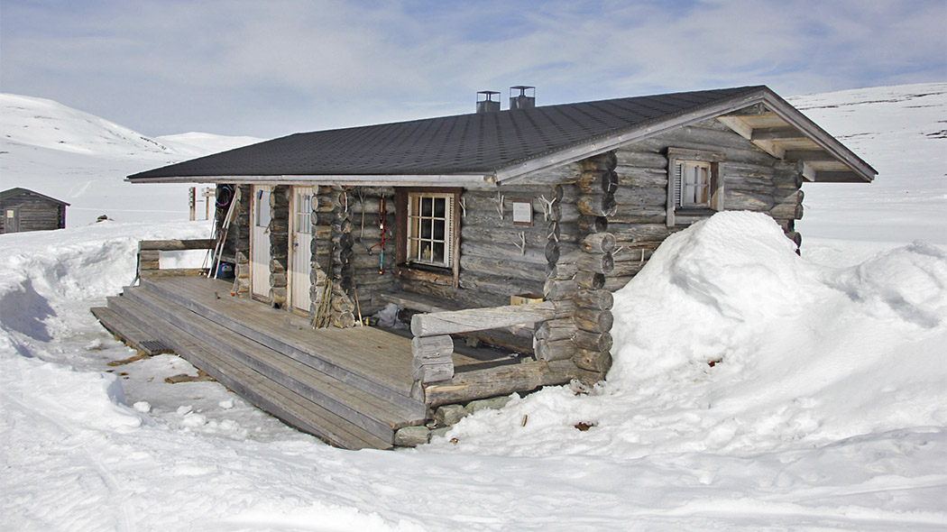 A log cabin in the middle of snow in open fell area with two doors and a full-length porch and stairs to the building.