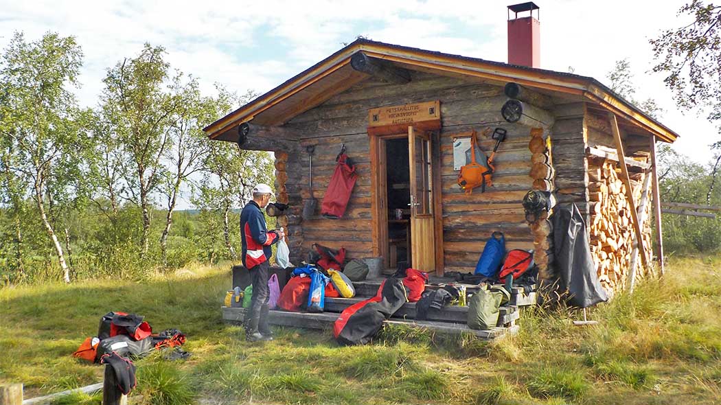 A log cabin in a summer fell. On the terrace of the cabin there are camping equipment and a man looking at the open door of the hut.