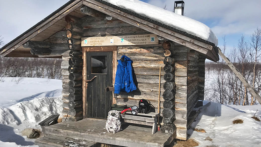 A log cabin with snow around and on the roof. There are camping equipment on the cabin´s terrace.