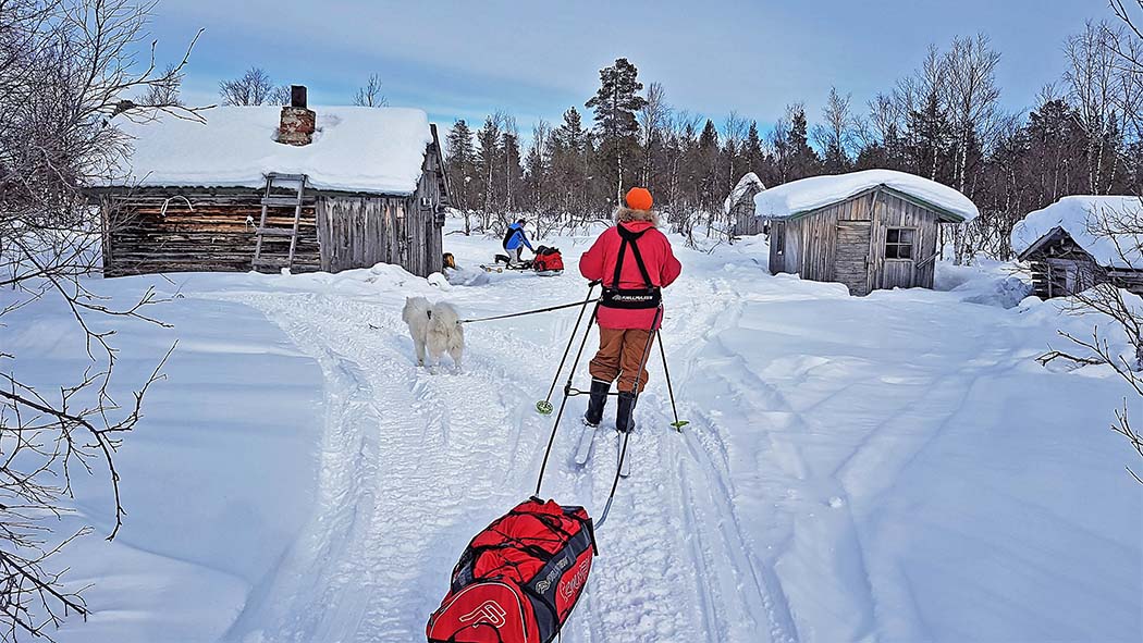 A skier pulling his equipment in pulk has arrived in the yard of the hut. Near the hut another hiker packs his gear.