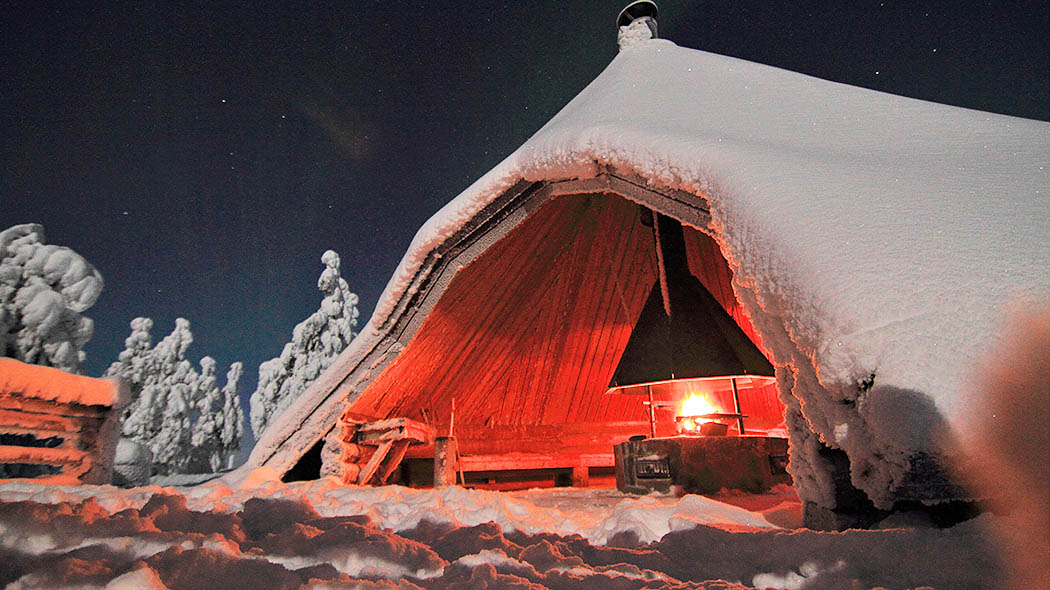 A large camfire shelter surrounded by snow-covered trees under the starry sky. A fire is burning in the hut. Above the fireplace is a conical flue.