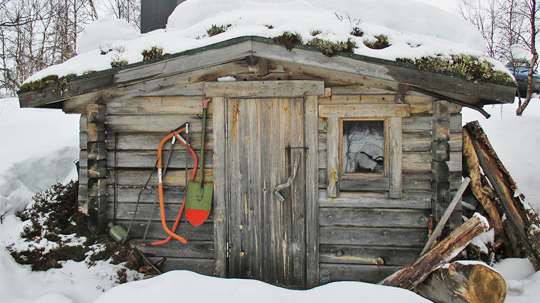 A small hut in a snowy landscape. There is a small window next to the door and a shovel and two saws hanging on the wall. The peat roof of the hut is almost completely covered with snow.