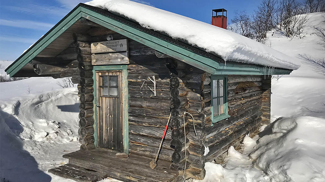 The log cabin is surrounded by snow in clear weather, and there is thick snow on the roof of the cabin. The door and window frames have been painted with turquoise.