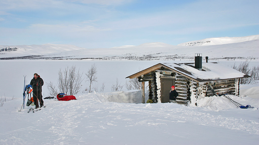 The log cabin is surrounded by snow. In the background there is a fell and a lake landscape. In front of the cabin there is a woman and a man with skis next to him.