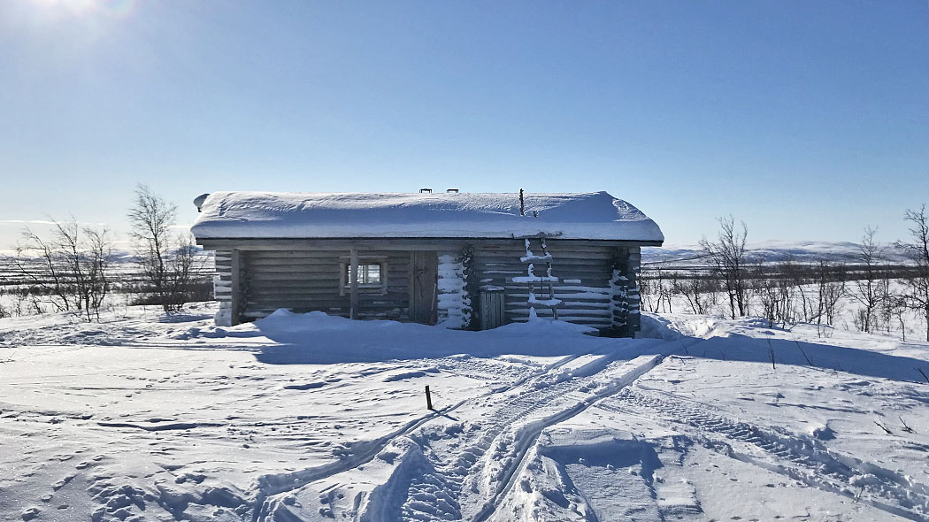Puuvrasjoki open wilderness hut on a sunny winter day. Snowmobile track marks can be seen on the snow.